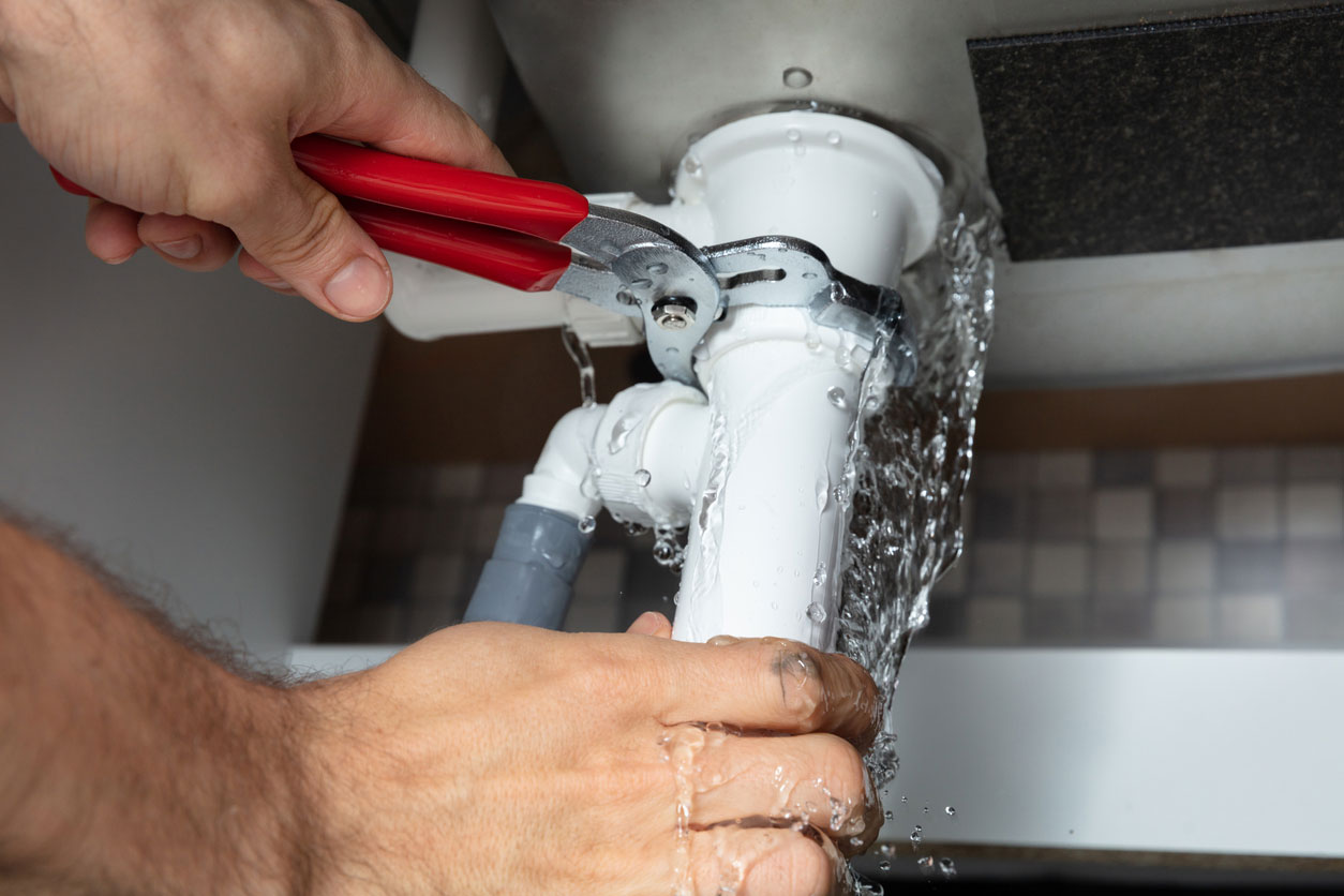 A plumber quickly repairs a leaking white pipe under a sink, water spraying as he works to contain the emergency.
